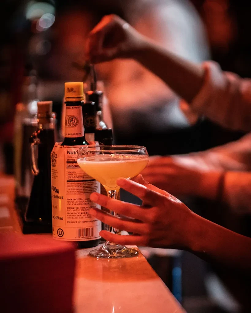 Close-up of a yellow cocktail, Angostura bitters, and hands at a dim bar.