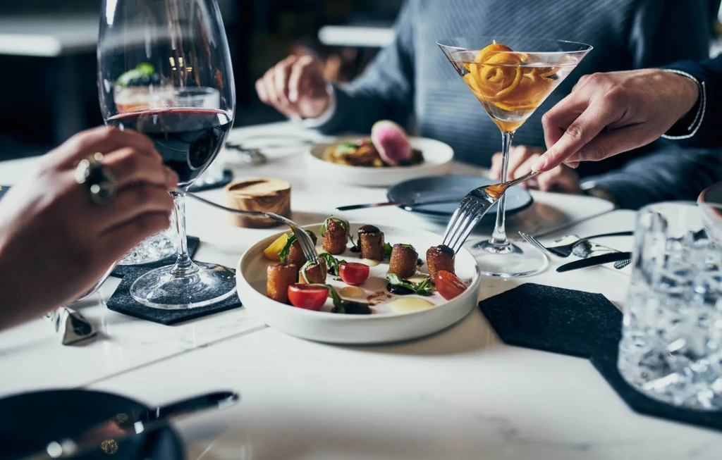Hands with forks over an appetizer, red wine, and a cocktail.