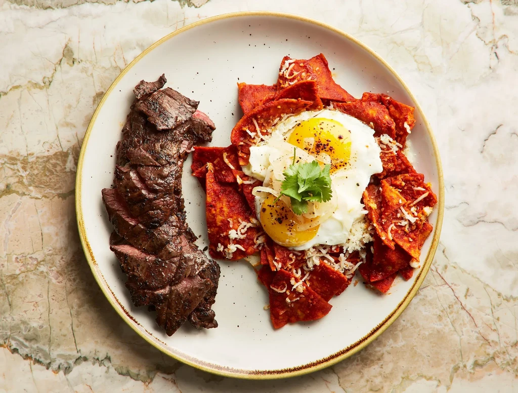 A plate of sliced grilled steak and red chilaquiles with fried eggs.