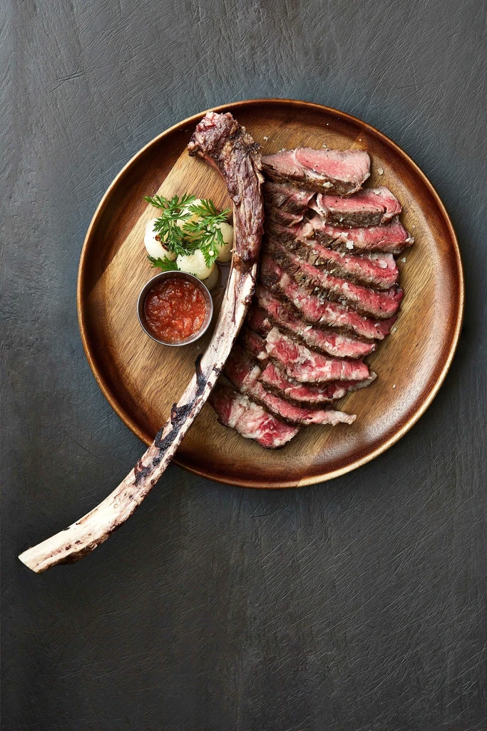 Sliced medium-rare tomahawk steak, roasted potatoes, red sauce, top-down view.