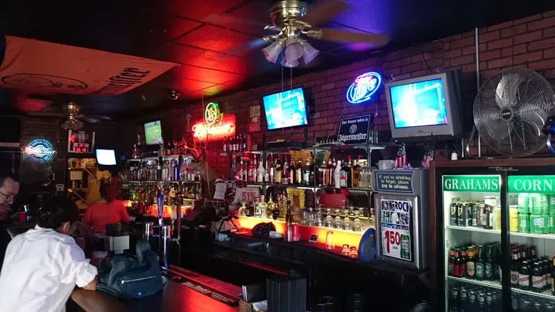 Dimly lit bar, neon signs illuminate bottles, TVs, and beer cooler.