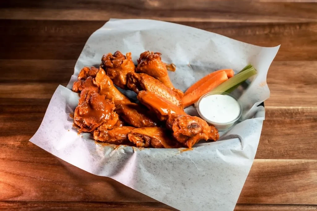 Glossy Buffalo chicken wings, carrots, celery, and dip in a basket.