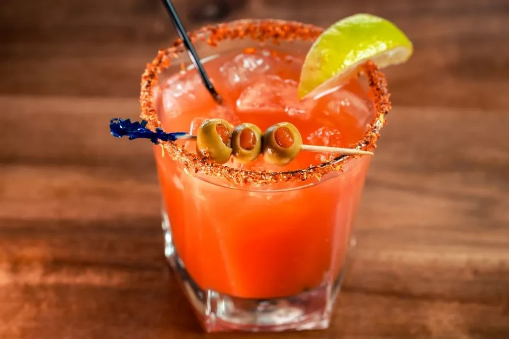 Red-orange cocktail, spiced rim, lime, and olive skewer in faceted glass.
