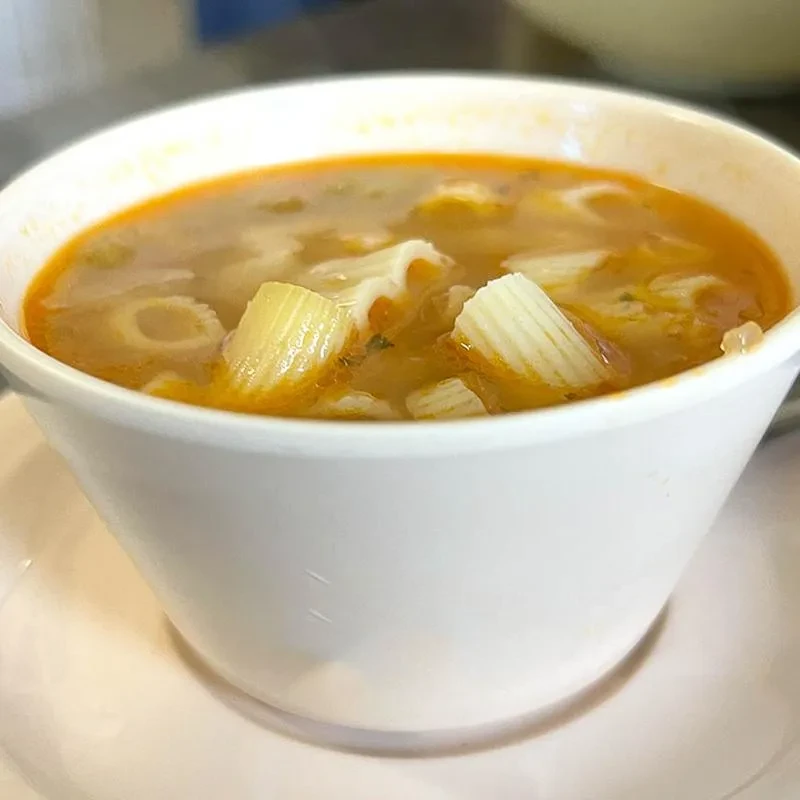 White bowl of orange-brown soup with tubular pasta and vegetables.