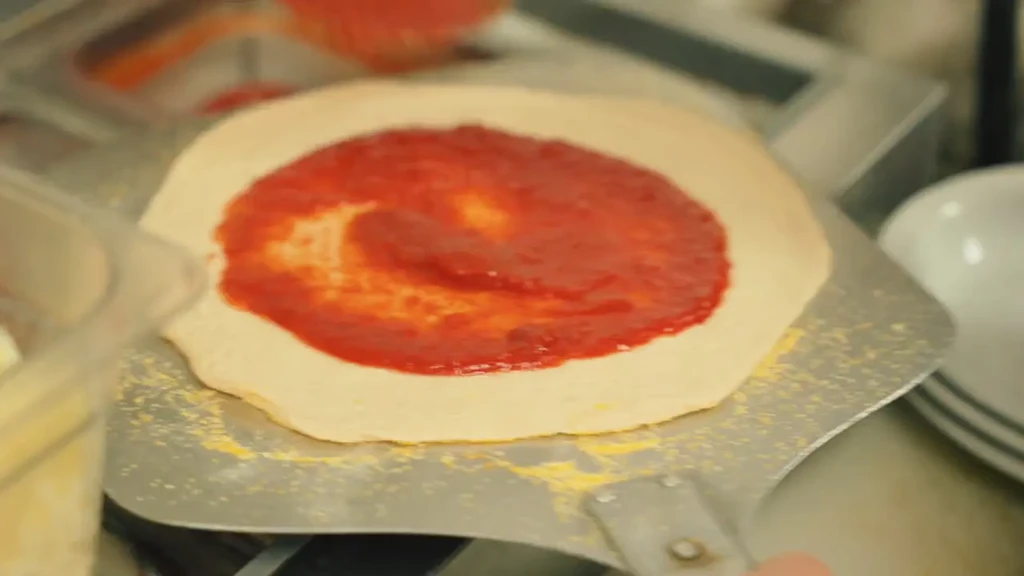 Raw pizza crust with tomato sauce on a metal pizza peel.