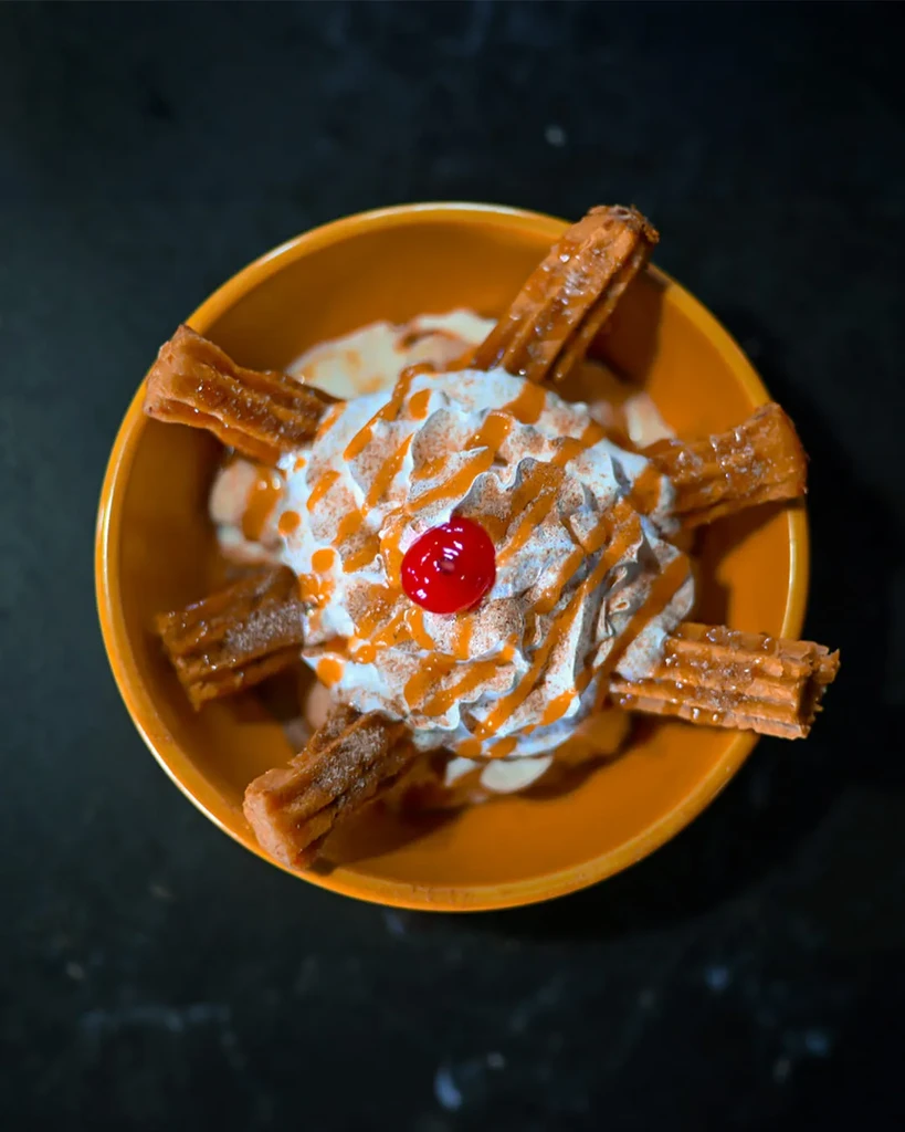 Golden churros with whipped cream, caramel, and cherry in an orange bowl.