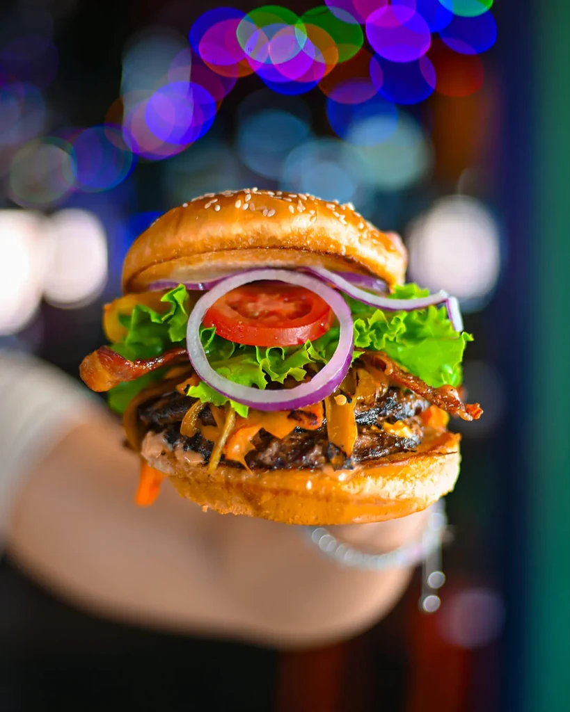 A person's hand holds a gourmet burger against colorful bokeh lights.