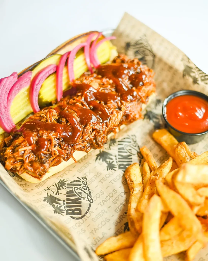 Saucy pulled pork sandwich, fries, and pickles on a metal tray.
