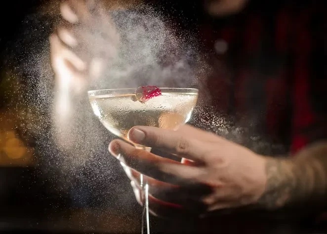 Close-up of golden cocktail in coupe glass, misted and held by tattooed hand.