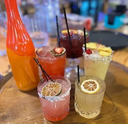 Five fruit-garnished cocktails and a Mango Soleia bottle on a bar.
