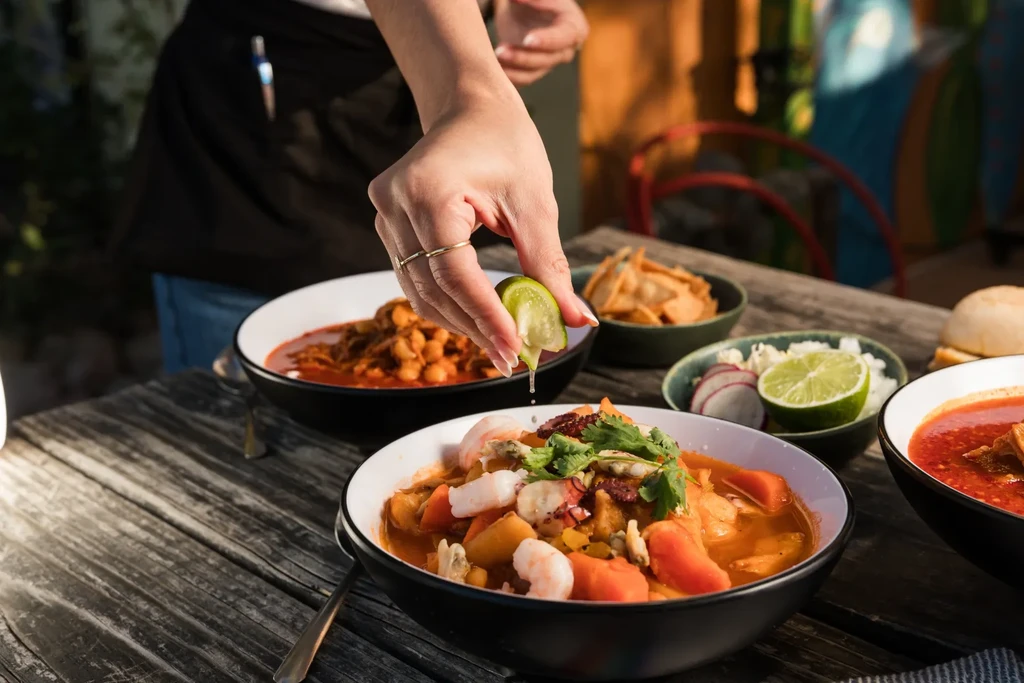 Hand squeezing lime over seafood stew, other dishes on a wooden table.