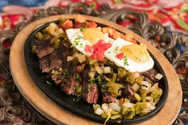 Grilled steak, fried eggs, and vegetables sizzling in a cast iron skillet.