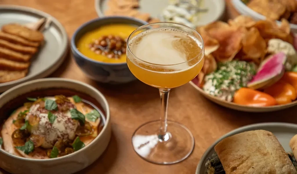An amber cocktail, soup, dips, crackers, and appetizers on a wooden table.