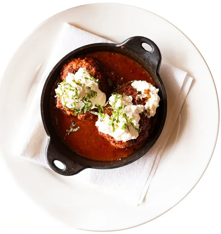 Two meatballs in a skillet with ricotta, sauce, and green herbs.