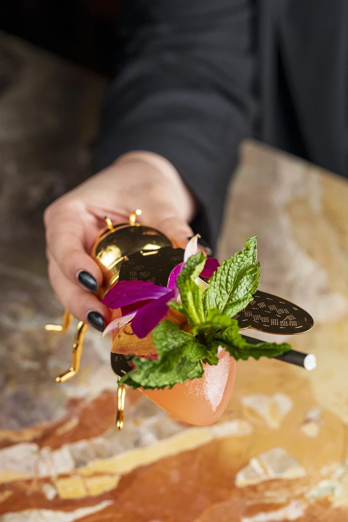 Golden beetle vessel holds coral cocktail with mint and orchid garnish.