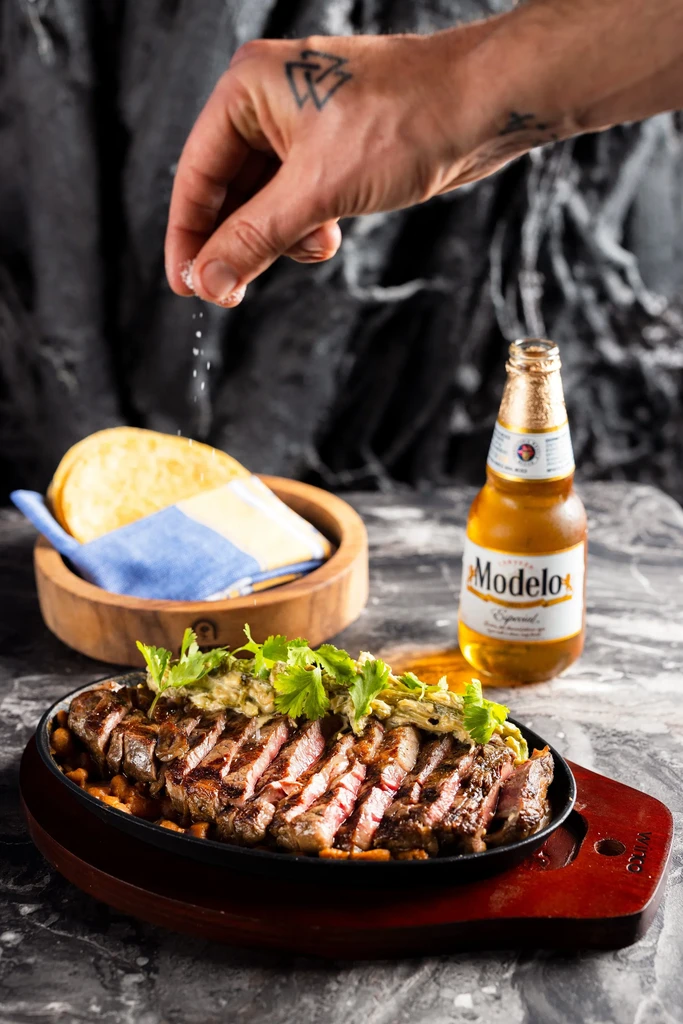 Tattooed hand salts sizzling sliced steak, with tortillas and Modelo beer.