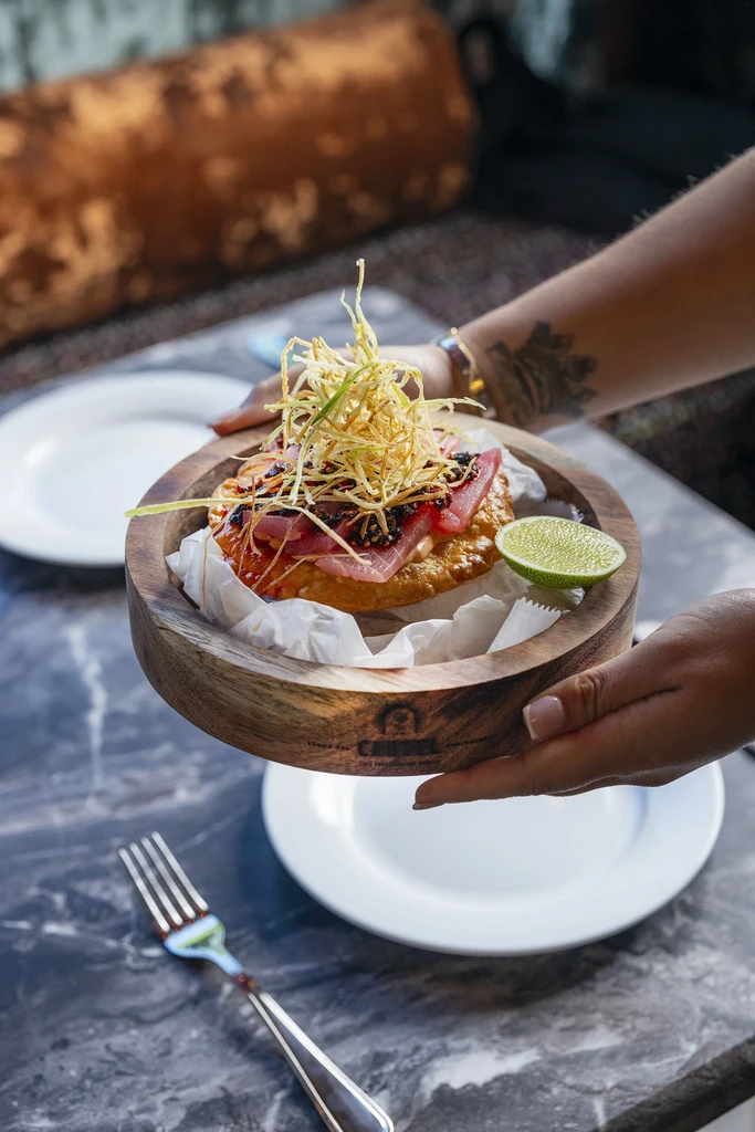 Hands present rare tuna dish with crispy topping in a wooden bowl.