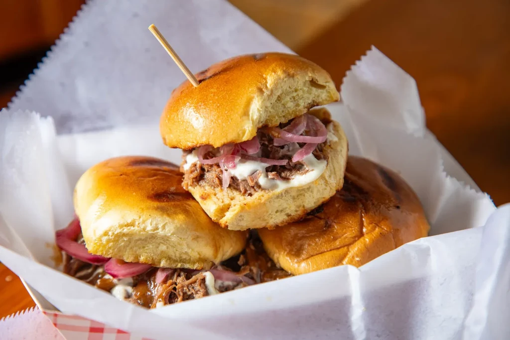 Three sliders in a basket, with shredded meat, sauce, and red onions.