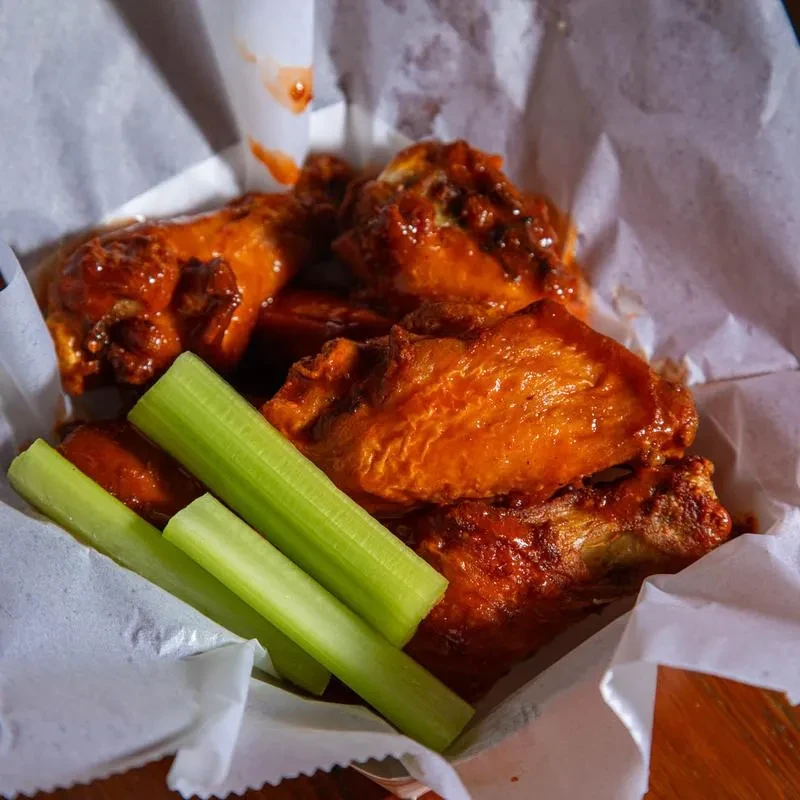 Reddish-orange buffalo wings with celery in a paper-lined basket.