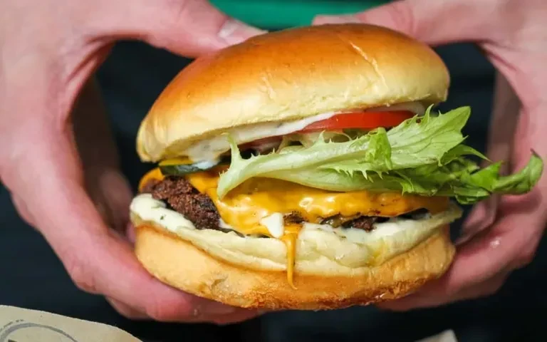 Hands holding a gourmet burger with cheese, vegetables, and dripping sauce.