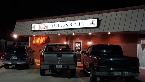 KJ's PLACE building at night, 'NOW OPEN' banner, parked vehicles.