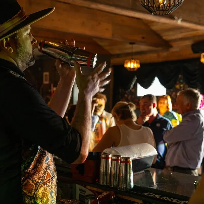 Male bartender in a hat shaking a cocktail shaker at a lively bar.