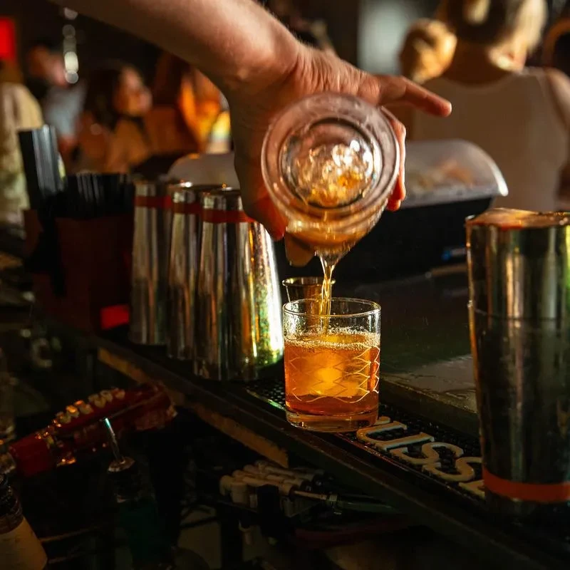 Bartender pours a stream of golden-brown liquid into an iced glass.