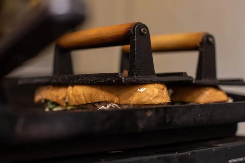 Two golden-brown sandwiches pressed on a grill by a cast-iron press.