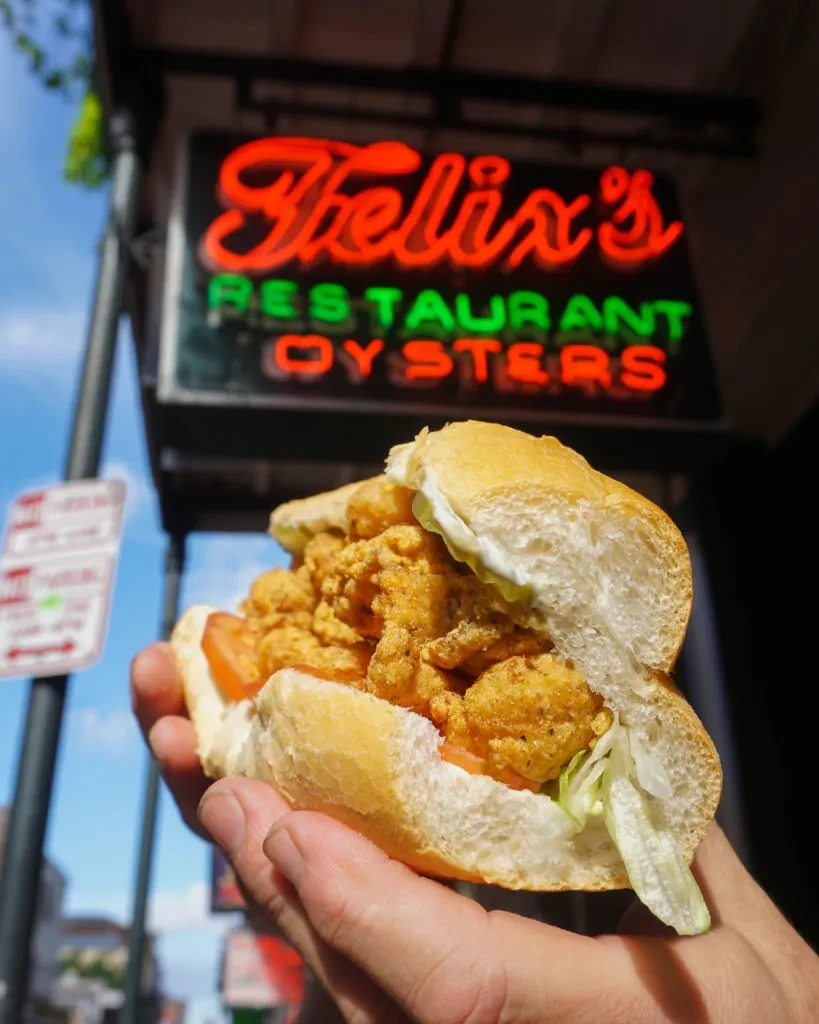 Hand holds a generously filled fried seafood po'boy near Felix's sign.