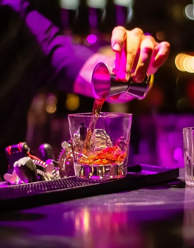 Hand pouring spirit into an ice-filled glass at a purple-lit bar.
