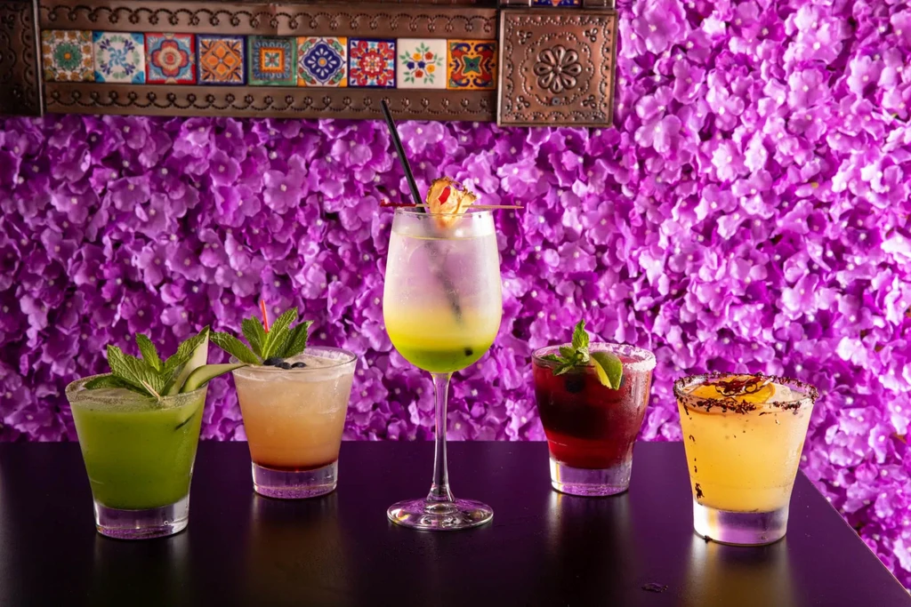 Five colorful cocktails with purple flower wall and patterned tile background.