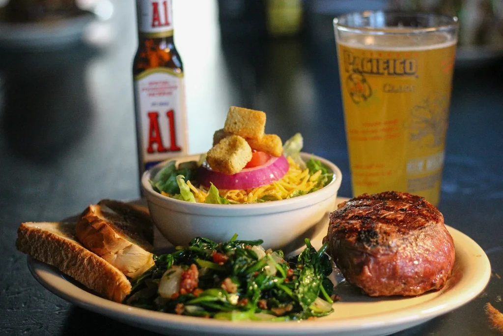 Medium-rare steak, greens, salad, toast, with A.1. sauce and beer.