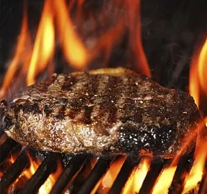 Thick meat patty with grill marks searing over flames.