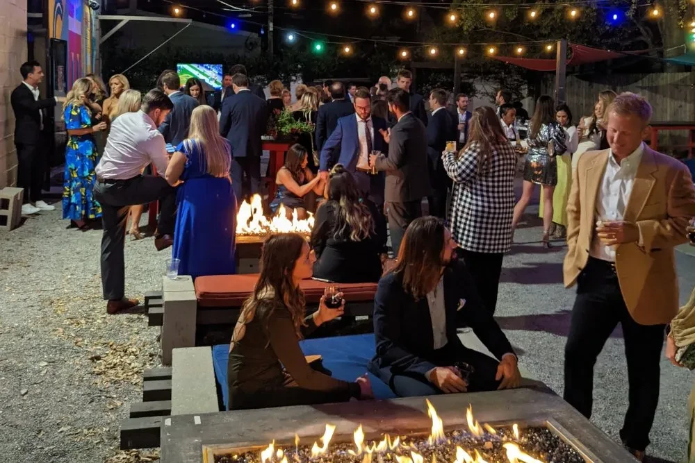 Guests mingle outdoors by fire pits under string lights at night.