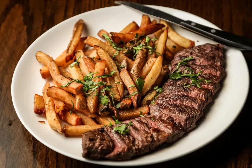 Close-up of steak frites with green herbs on a dark table.