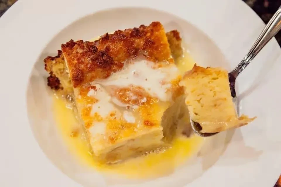 Spoon scoops golden-brown bread pudding with creamy white and buttery sauces.
