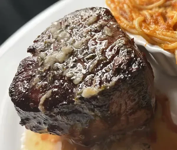 Thick seared steak topped with garlic-herb butter; pasta in blurred background.