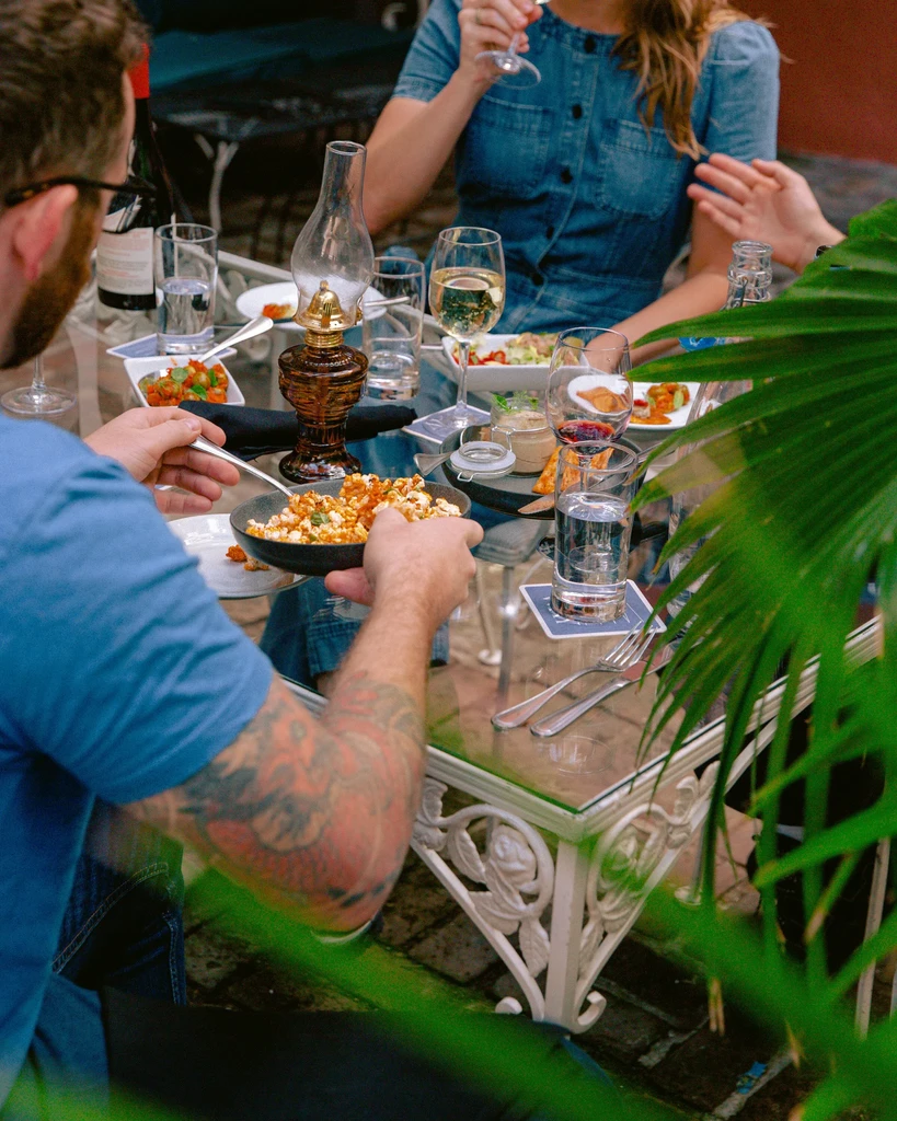 Outdoor dining table with food, wine, and three partial diners.