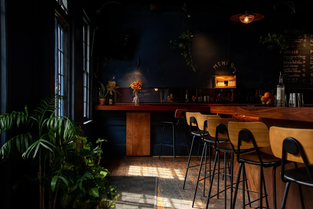Dimly lit bar interior with long wooden counter, stools, and chalkboard menu.