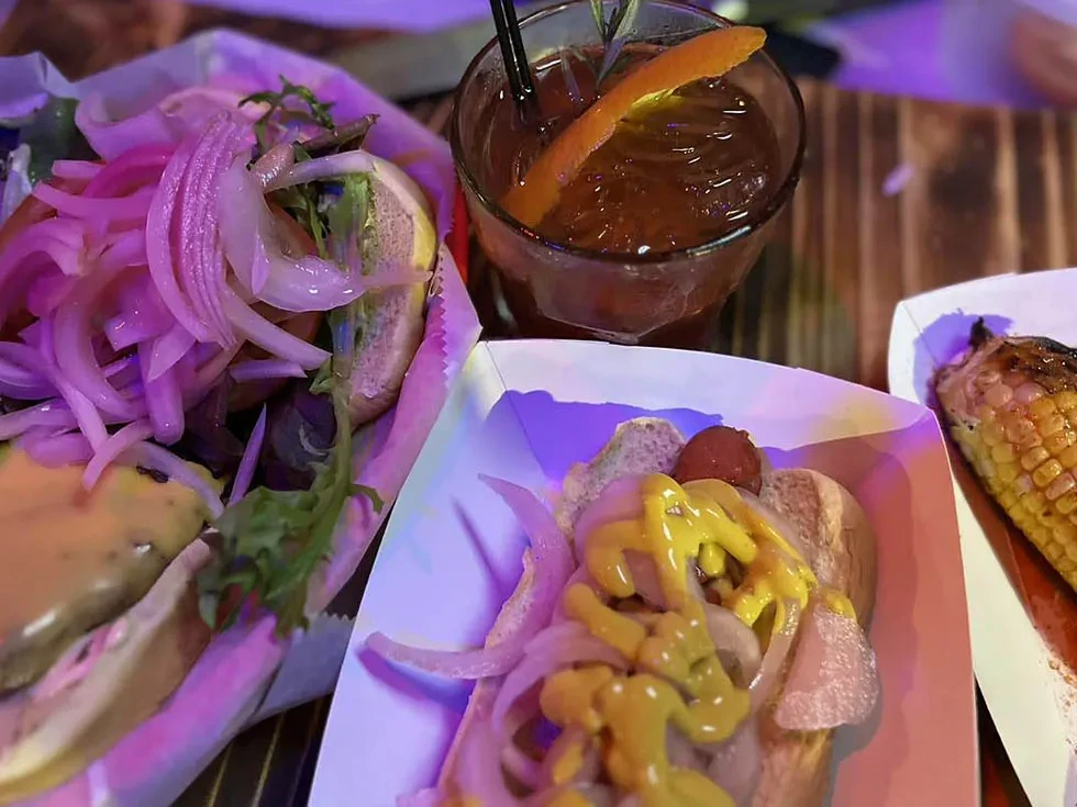 Burger, hot dog, corn, and cocktail in purple-pink ambient light.