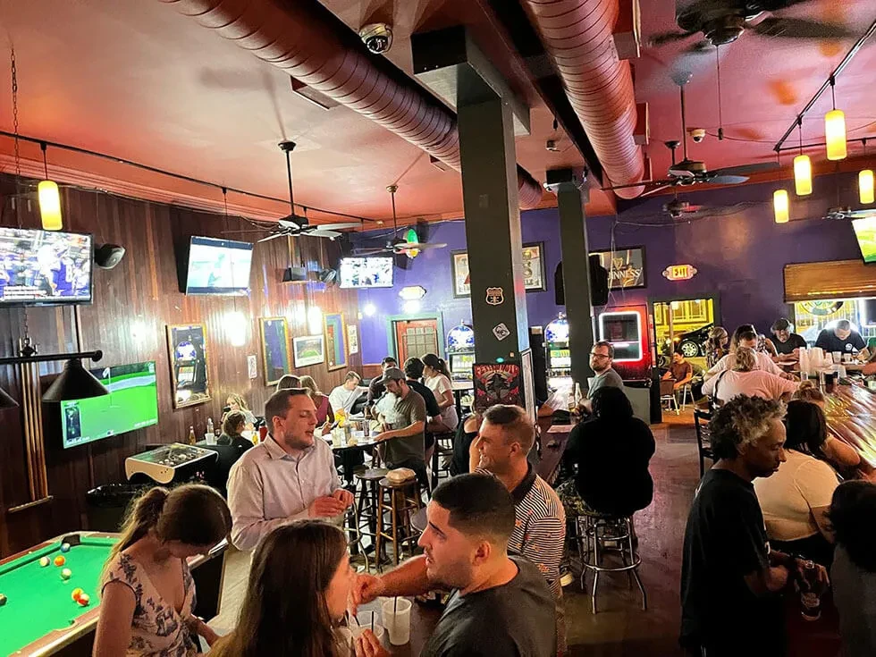 Bustling bar interior with patrons playing pool and watching sports on TVs.