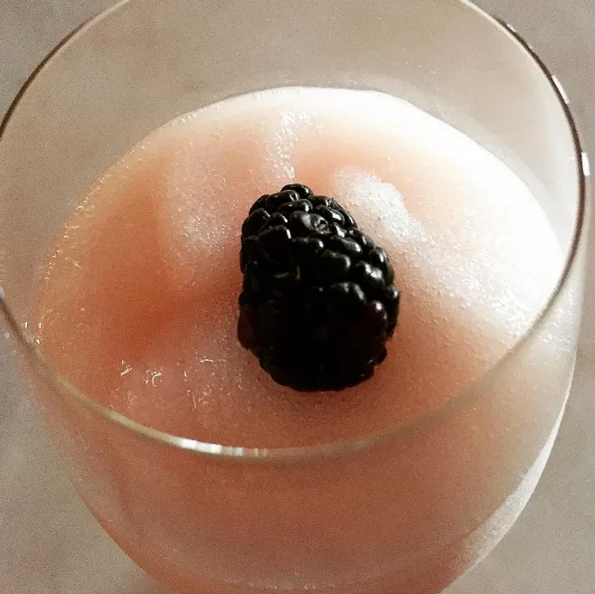 Light pink frothy drink in a stemmed glass, topped with a blackberry.