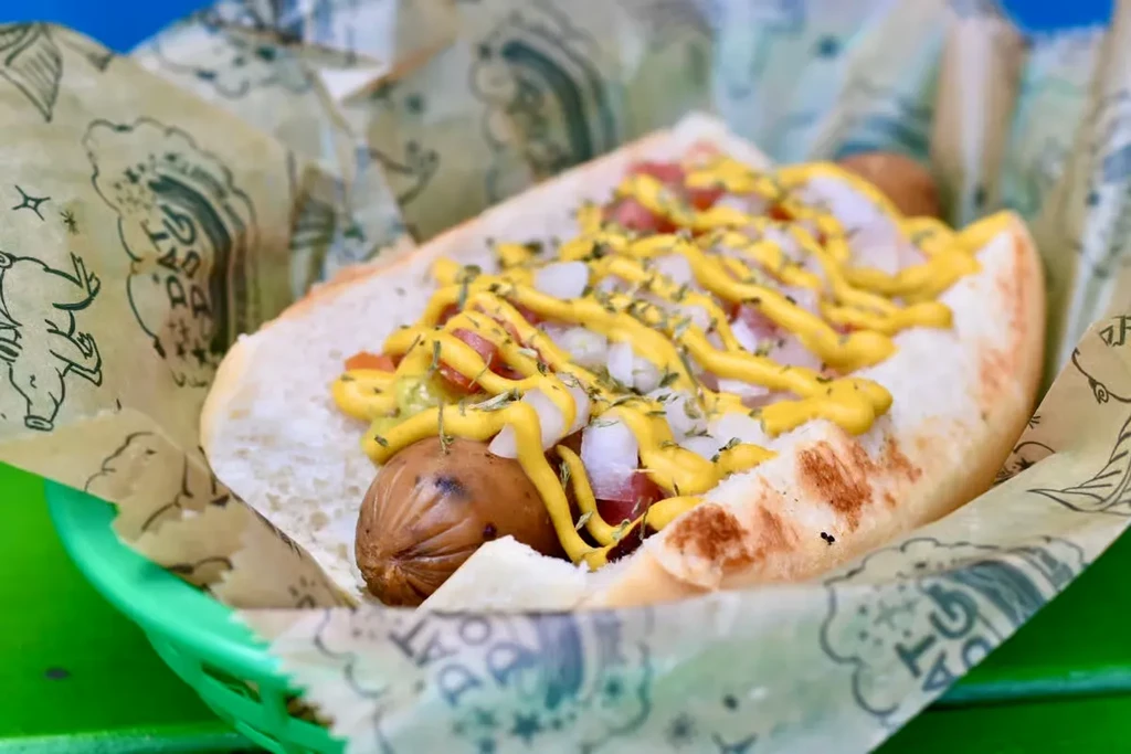 Grilled hot dog with mustard, onions, tomatoes, and herbs in a pig-patterned basket.