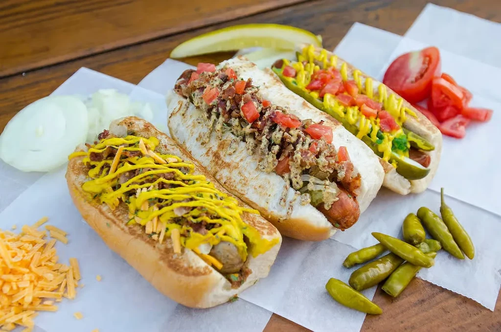 Three gourmet hot dogs with diverse toppings and fresh accompaniments on wood.