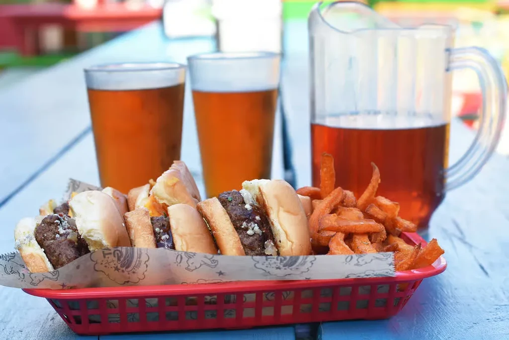 Close-up of sliders, sweet potato fries, and amber drinks outdoors.