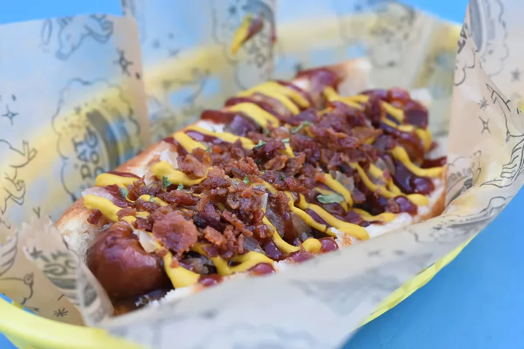 Close-up hot dog topped with onions, bacon, mustard, reddish sauce, green herbs.