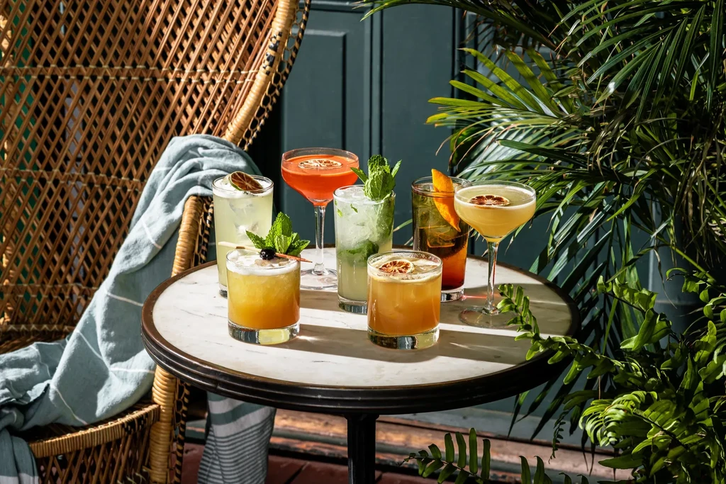 Seven vibrant cocktails on a marble table with tropical plants.