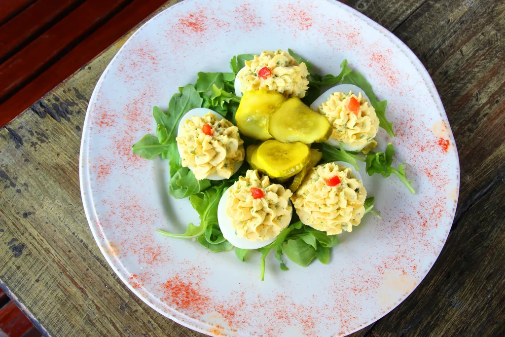 Six garnished deviled eggs with arugula and pickles on a wood table.