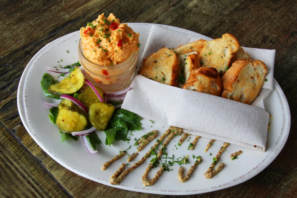 Appetizer plate with orange dip, pickles, toast, and mustard on wood.
