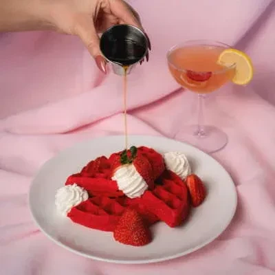Hand pouring syrup on red waffles with cream and strawberries, pink cocktail.